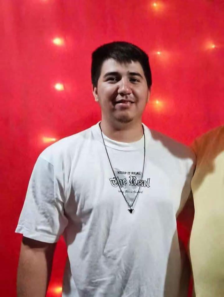Aldo Alexis Duarte sonriente, en camiseta blanca con el texto 'Keep it Real', fondo rojo decorado con luces.