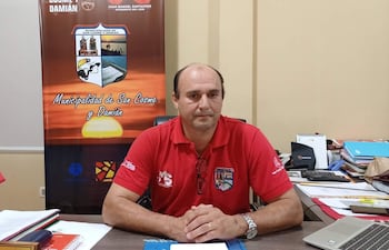 Juan Manuel Santacruz vestido con camiseta roja sentado en un escritorio, con documentos y una computadora en su oficina, luciendo tranquilo.