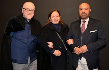 Hombre canoso con gafas y capa negra, mujer con abrigo negro y hombre calvo con chaleco y corbata, todos sonriendo en un entorno oscuro.