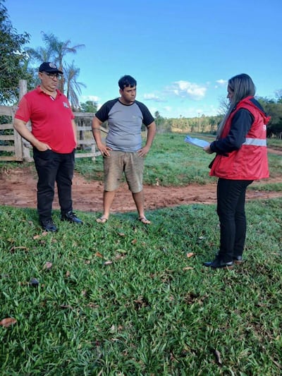 Mujer con chaqueta roja sostiene un documento, conversando con dos hombres en un ambiente natural y tranquilo.