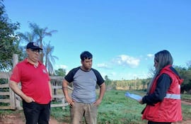 Mujer con chaqueta roja sostiene un documento, conversando con dos hombres en un ambiente natural y tranquilo.