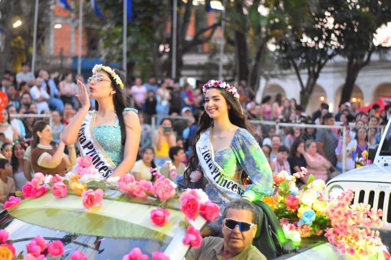 Jóvenes con coronas de flores y vestidos brillantes en vehículo decorado, mientras un hombre mayor al volante sonríe.