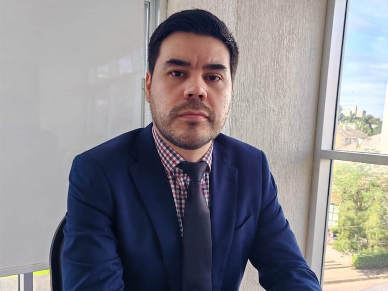 Ricardo Estigarribia sentado en una mesa, vestido con blazer azul y corbata oscura, con expresión tranquila en un entorno iluminado.