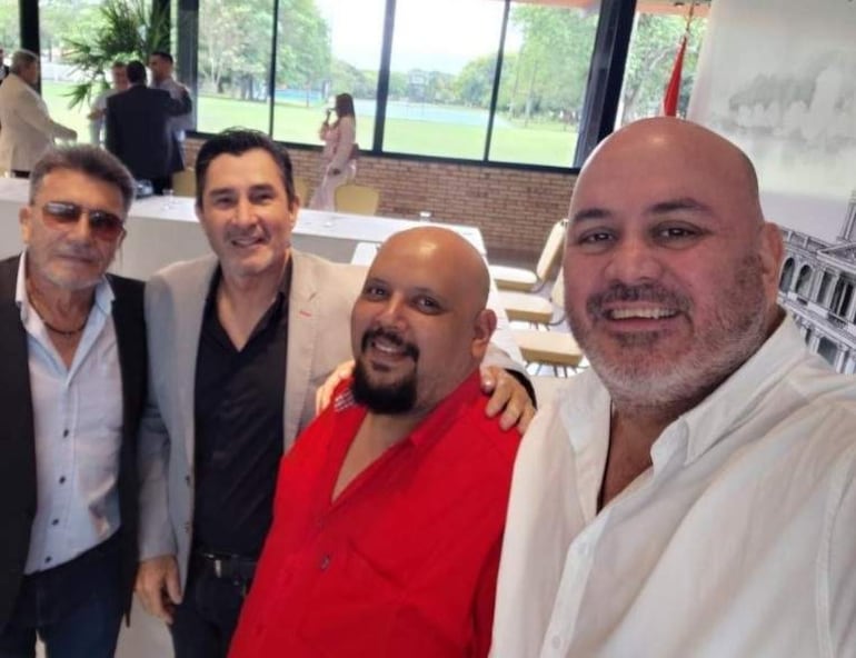 Cuatro hombres sonrientes: uno con gafas en traje oscuro, otro en chaqueta clara, un tercero en camiseta roja y el último en camisa blanca.