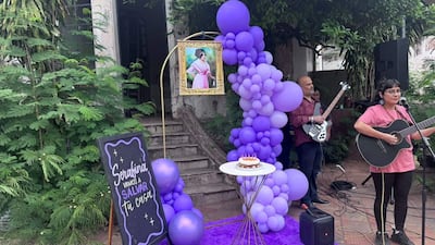 Mujer cantando con guitarra y hombre en segundo plano tocando el bajo, entre globos morados y un cartel que dice 'Serafina vamos a salvar tu casa'.