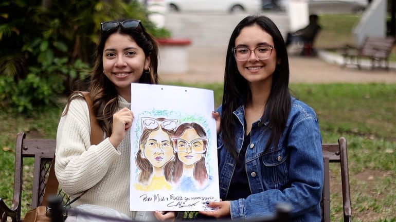 Dos mujeres sonriendo en un parque, una con suéter blanco y otra con chaqueta de mezclilla sosteniendo un colorido retrato.
