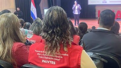Mujer con chaqueta roja en auditorio, mientras otra habla desde un podio en un evento sobre protección infantil.