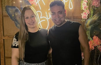 Mujer con camiseta negra y falda de mezclilla, y hombre con chaleco sin mangas, sonriendo en una celebración de cumpleaños decorada.