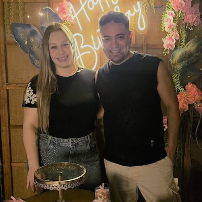 Mujer con camiseta negra y falda de mezclilla, y hombre con chaleco sin mangas, sonriendo en una celebración de cumpleaños decorada.