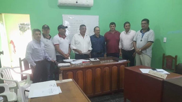 Siete hombres de pie en sala con paredes verdes, discutiendo documentos en mesa, todos con expresiones tranquilas.