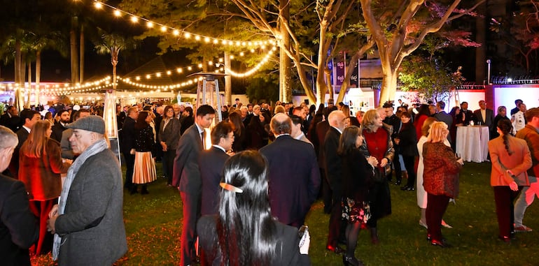 Grupo de personas conversando y disfrutando de música en un jardín iluminado, con ambiente festivo y elegante.