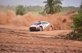Automóvil de rally en acción, levantando polvo en un terreno rural polvoriento con vegetación al fondo.