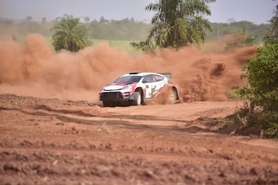 Automóvil de rally en acción, levantando polvo en un terreno rural polvoriento con vegetación al fondo.