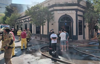 Grupo de bomberos en indumentaria especializada, manipulando mangueras, junto a civiles preocupados frente a la discoteca Nápoles.