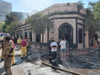 Grupo de bomberos en indumentaria especializada, manipulando mangueras, junto a civiles preocupados frente a la discoteca Nápoles.