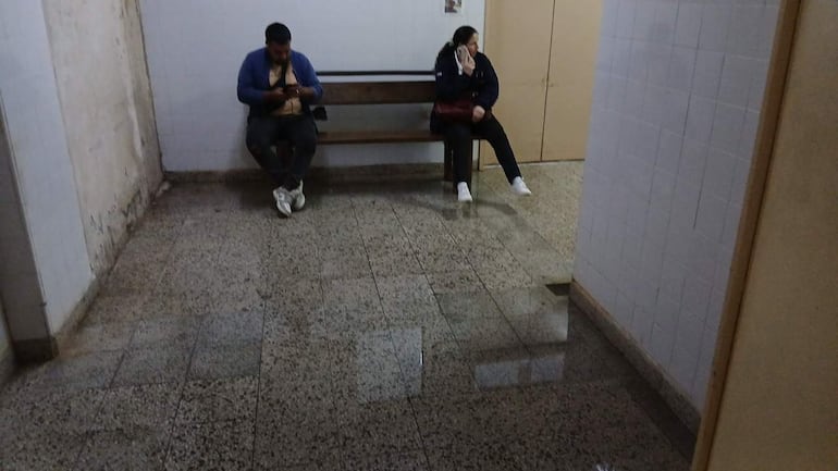 Hombre con suéter azul y mujer con abrigo oscuro se sientan en un banco de madera, concentrados en sus celulares.