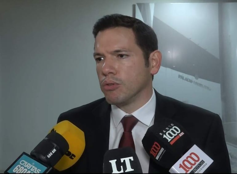 Alejandro Aguilera, vestido con traje oscuro y corbata, habla serio rodeado de micrófonos de prensa en un entorno formal.