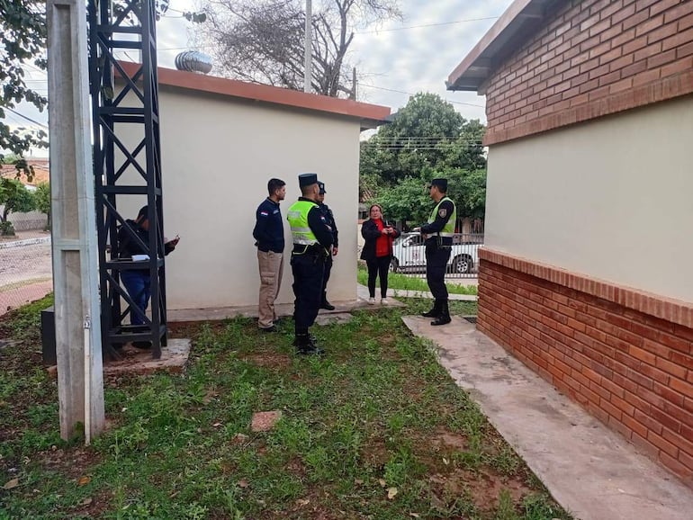 Cuatro personas, incluidos dos policías en uniforme, atienden a una mujer de chaqueta roja en un entorno verde cerca de edificios de ladrillo.