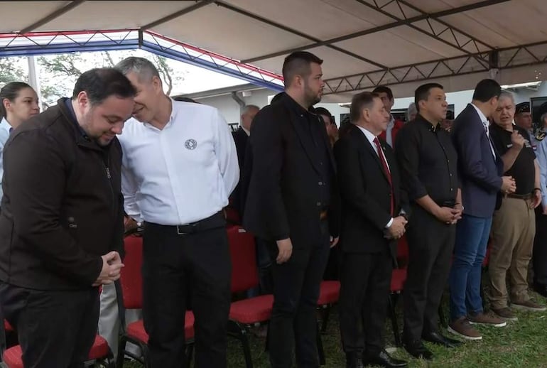 Hugui Farías con traje oscuro y Carlos Benítez conversan en un evento formal bajo una carpa.