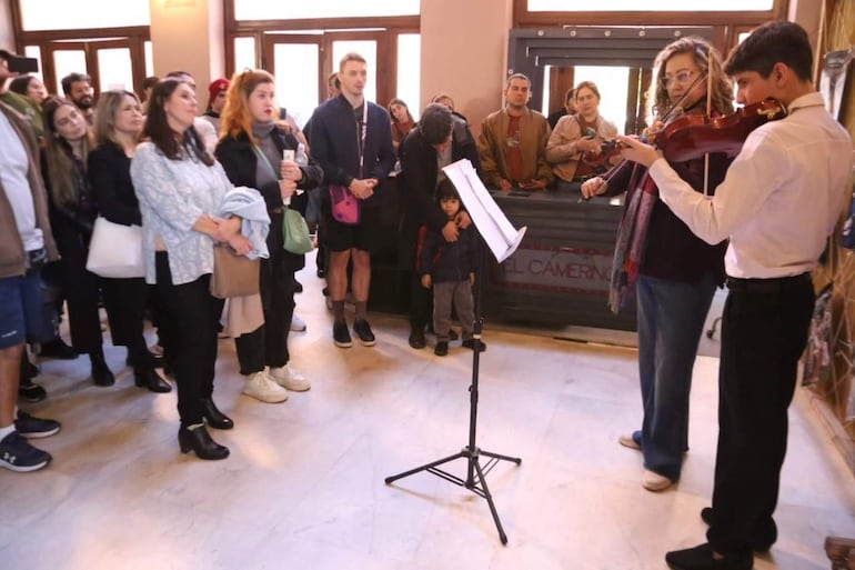 Mujer con gafas y niño en chaqueta oscura tocan violín en un escenario iluminado, mientras adultos observan atentos.