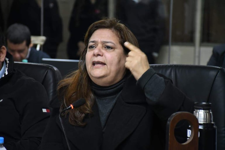 Rossana Rolon gesticula en chaqueta oscura, mientras otros participantes la observan en un ambiente formal.