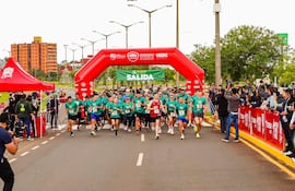 Multitud de corredores con camisetas verdes y rojas en la salida de la carrera, bajo un arco inflable. Ambiente urbano de fondo.
