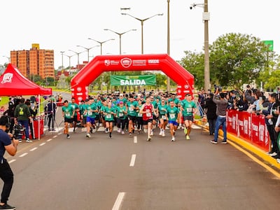 Multitud de corredores con camisetas verdes y rojas en la salida de la carrera, bajo un arco inflable. Ambiente urbano de fondo.