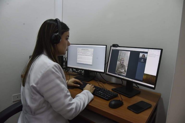 Mujer con bata blanca y auriculares frente a computadora, asistiendo a otra mujer en videollamada desde un consultorio.