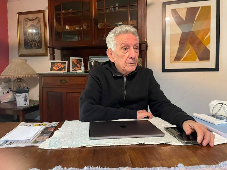 Hombre canoso, con chaqueta oscura, sentado en mesa con ordenador portátil y documentos, expresión pensativa en ambiente acogedor.