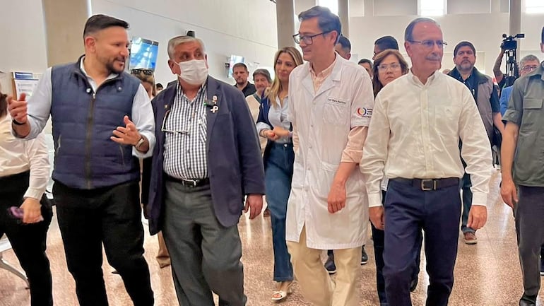 Isaías Fretes en bata blanca, rodeado de bioquímicos en un ambiente formal, discutiendo con expresiones serias y tristes.