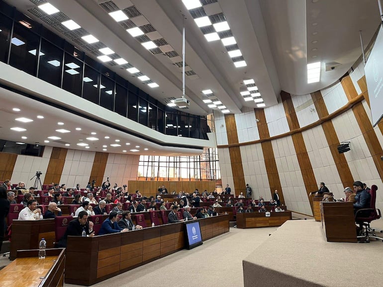 Asistentes en la audiencia pública con expresión tranquila, en un salón moderno con luz brillante y grandes ventanales.