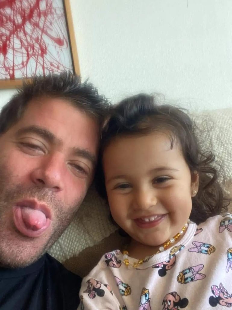 Niña pequeña sonriendo, posando para una selfie con un collar colorido. Fondo desenfocado.