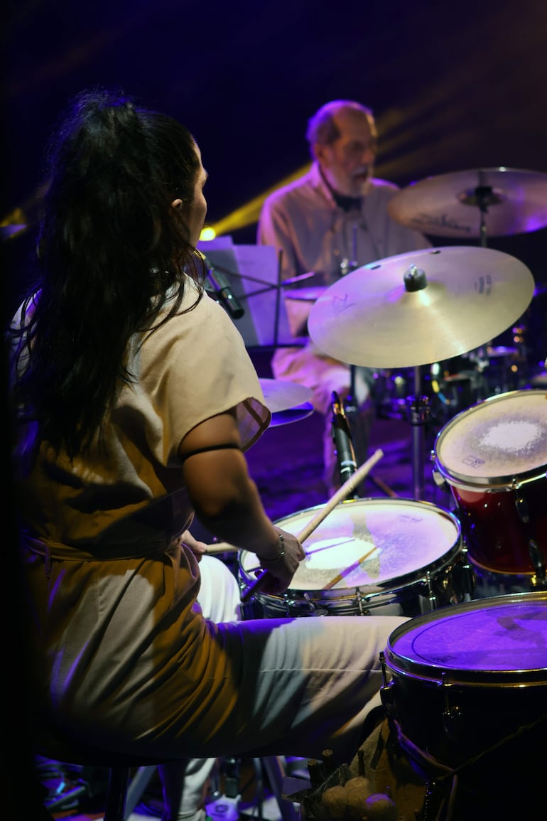 Mujer con cabello largo y oscuro en camiseta clara toca la batería, mientras hombre con barba toca un instrumento de percusión.