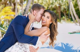 Leah Ashmore con vestido blanco de encaje, abrazada por un hombre con traje azul oscuro, ambos sonrientes en un entorno de vegetación tropical.