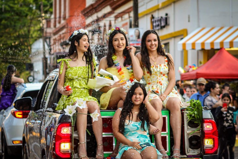 Cuatro mujeres sonriendo en la parte trasera de una camioneta, vestidas con colores vibrantes, celebran con flores en un ambiente festivo.