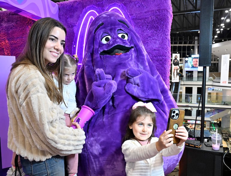 Mujer con suéter suave sonríe sosteniendo a una niña con lazo rosa, junto a figura disfrazada de Grimace en ambiente festivo.