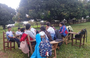 Jóvenes sentados en sillas de madera en un parque, algunos escuchando y otros participando en una conversación, rodeados de césped y árboles.