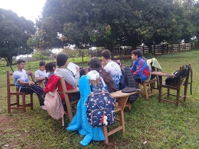 Jóvenes sentados en sillas de madera en un parque, algunos escuchando y otros participando en una conversación, rodeados de césped y árboles.