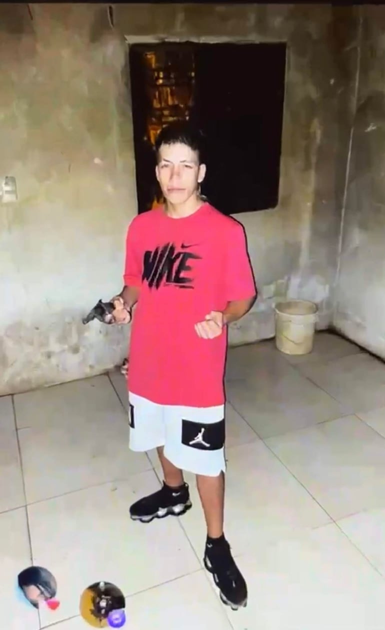 Joven con camiseta roja de Nike y pantalones cortos blancos sostiene un arma en un ambiente poco iluminado, con expresión seria.