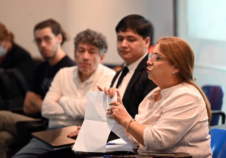 Mujer con blusa blanca y gafas presenta en auditorio, rodeada de tres hombres escuchando con atención.