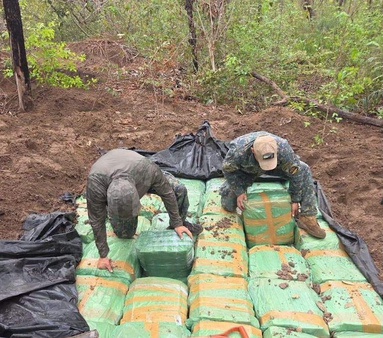 Dos hombres agachados en un entorno natural, uno con abrigo de camuflaje y otro en uniforme militar, manipulando paquetes envueltos.