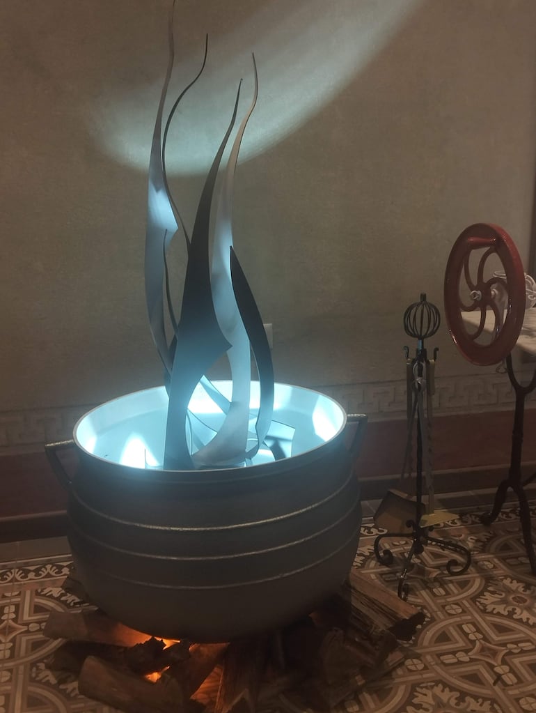 Instalación artística de un caldero con figuras metálicas iluminadas en azul, sobre troncos, en un ambiente interior moderno.