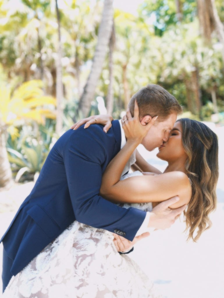 Hombre en traje azul oscuro y mujer en vestido blanco floral se besan, rodeados de vegetación tropical.