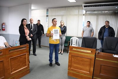 Hombre con camiseta amarilla sostiene documento con el texto 'EXPEDIENTE MUNICIPAL TRABADO', mientras varias personas lo observan en sala.
