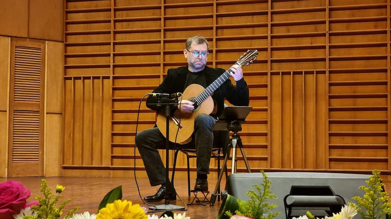 José Antonio Escobar, vestido con jeans oscuros y saco negro, toca guitarra con expresión concentrada en un ambiente con flores decorativas.