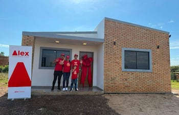 Cuatro personas en camisetas rojas y gorras con el logo 'Alex' celebran frente a una casa nueva, sosteniendo regalos.
