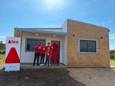 Cuatro personas en camisetas rojas y gorras con el logo 'Alex' celebran frente a una casa nueva, sosteniendo regalos.
