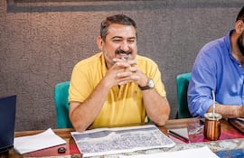 Luis Yd sonríe en camiseta amarilla, sentado frente a otro hombre de camisa azul en una mesa moderna con documentos y un portátil.