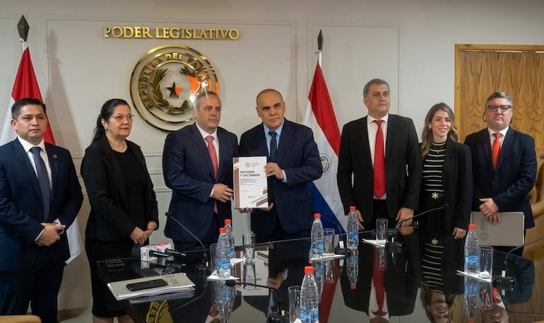 Seis personas en sala del Congreso, dos sostienen documento, banderas paraguayas visibles, ambiente formal y serio.
