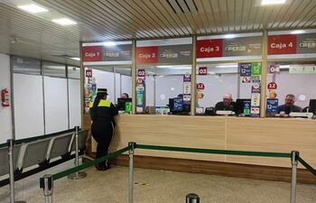 Policía en uniforme consulta en Caja 2, mientras tres personas mayores esperan conectadas a sus tareas, el entorno destaca por su iluminación.